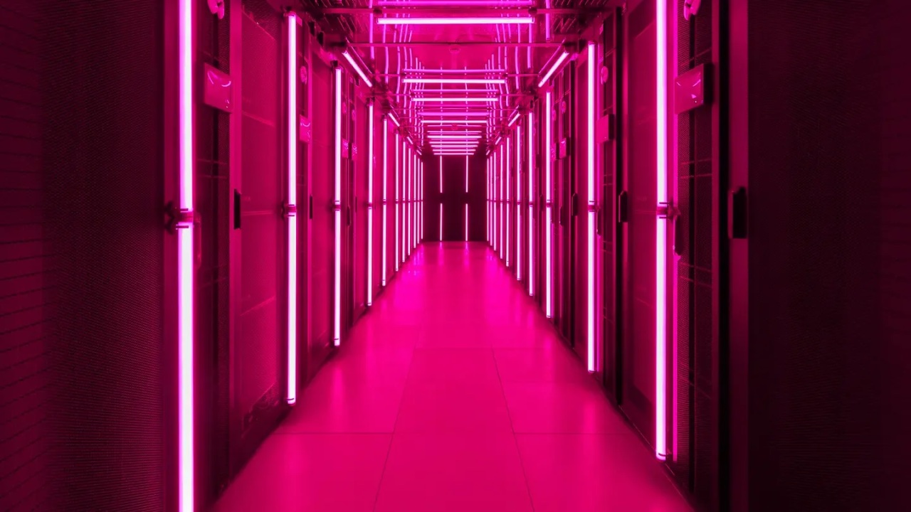 A long data-center corridor lined with server cabinets, lit by bright magenta neon tubes that recede to a distant vanishing point; the polished floor glows pink.